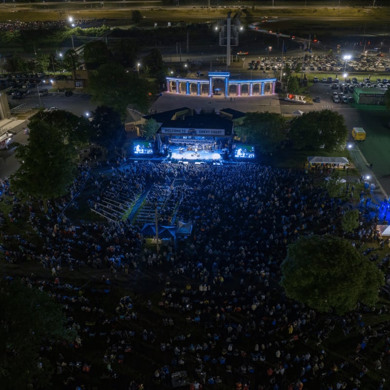 A large crowd gathers in the foreground before a brightly lit outdoor stage, with a glowing blue building and a parking lot in the background.