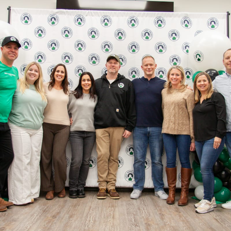 A group of people stand together in the foreground against a white backdrop featuring a repeating circular logo and green balloons.