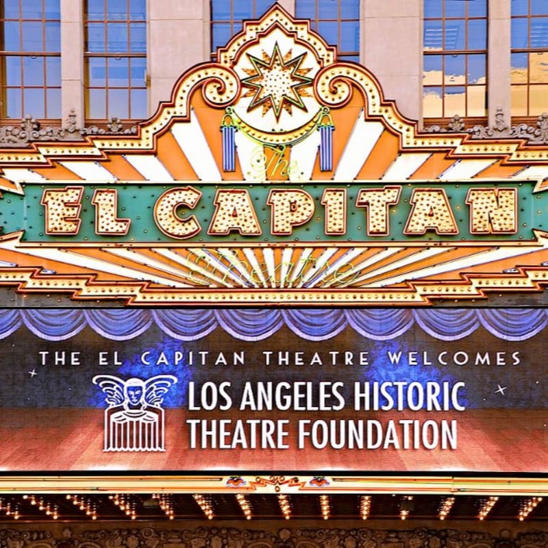 An ornate, illuminated El Capitan Theatre marquee stands in the foreground against a backdrop of a building with blue-tinted windows.