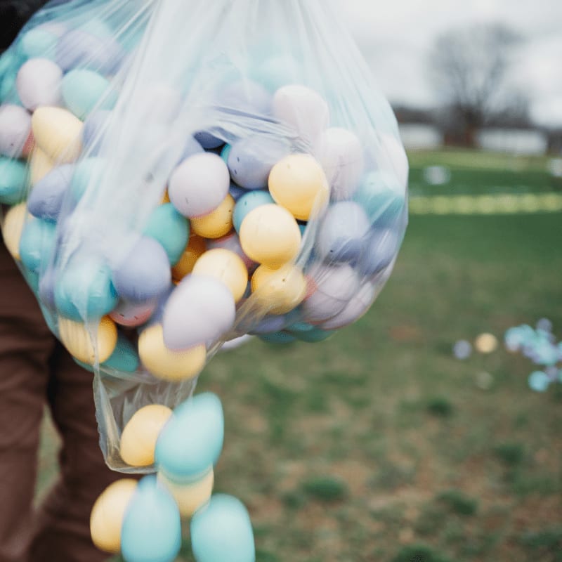 A clear plastic bag filled with colorful pastel Easter eggs is held in the foreground against a blurred green grassy field.