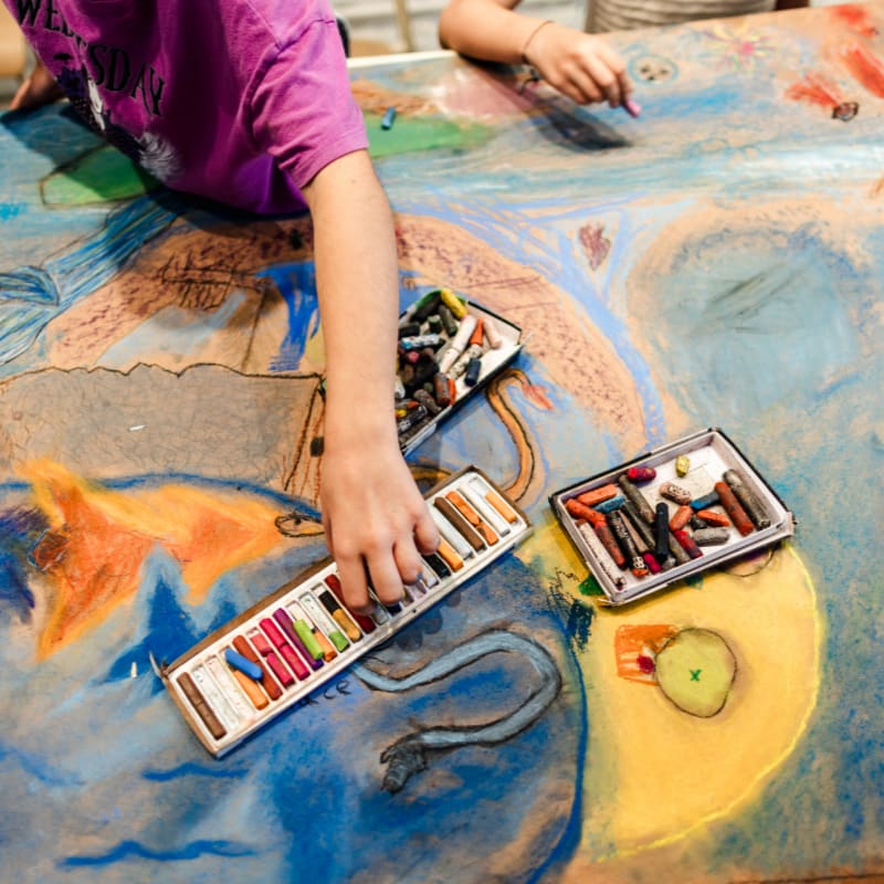 A person's hand reaches for a box of colorful pastels on a large, vibrant piece of art, with another person drawing in the background.