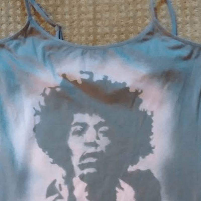 A blue tank top featuring a graphic print of Jimi Hendrix is laid against a textured beige background.