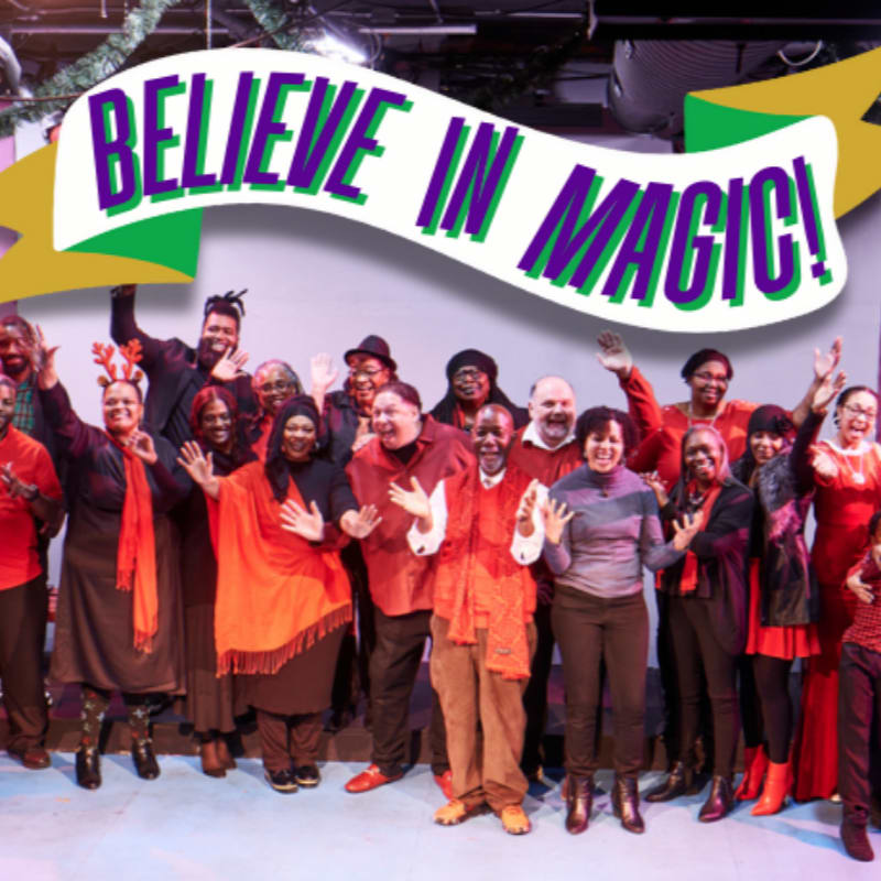 A diverse group of people in red and black attire pose together under a large "BELIEVE IN MAGIC!" banner against a plain light-colored background.