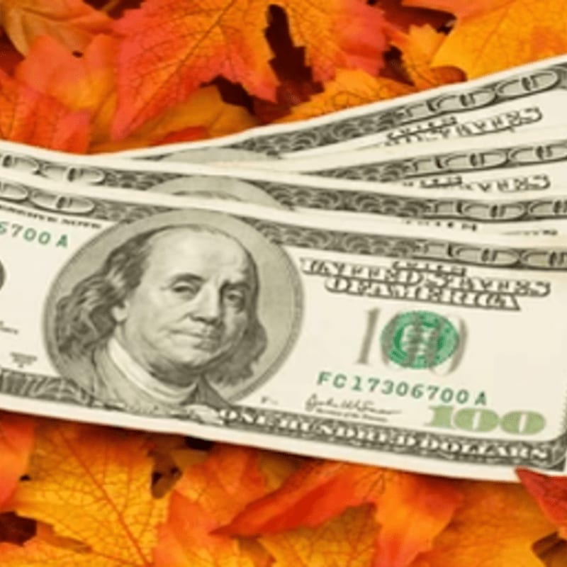 Several one-hundred-dollar bills lie scattered across a bed of vibrant orange and red autumn leaves.