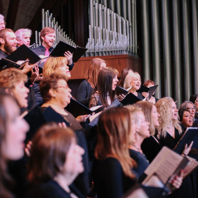 A choir of singers in dark clothing holds sheet music in the foreground, with large organ pipes rising in the background.