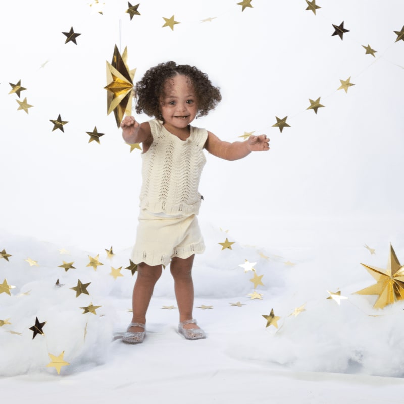 A smiling toddler in a cream-colored outfit stands amidst fluffy white clouds and gold stars in the foreground and background.