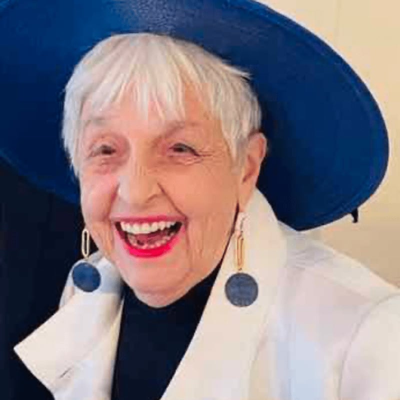 A smiling elderly woman with white hair, wearing a large blue hat and white blazer, stands against a plain, light-colored background.