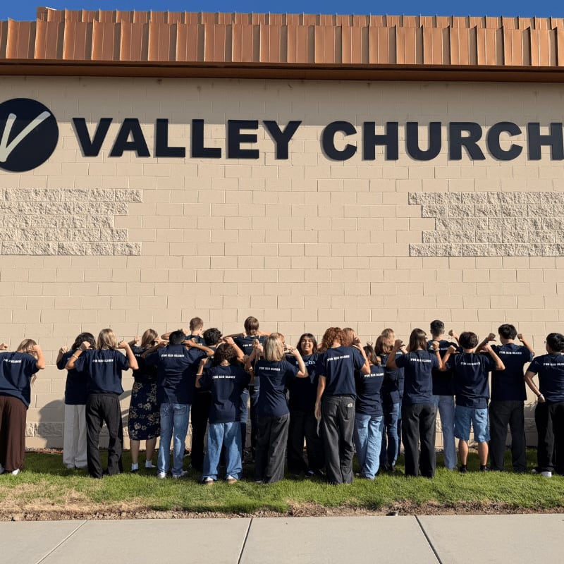 A group of people stand in a circle on a grassy area in front of a tan building with "VALLEY CHURCH" written on the wall.