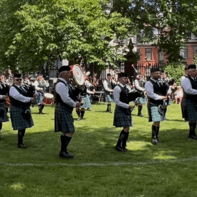 A group of bagpipers in traditional kilts perform on a grassy lawn surrounded by lush green trees and brick buildings.