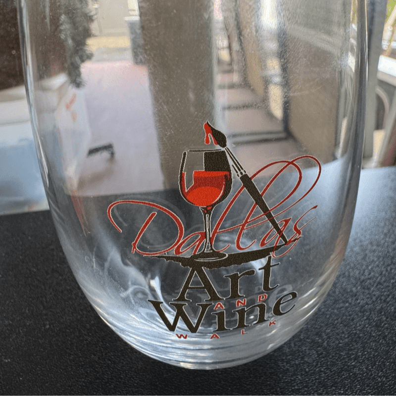 A clear glass with a "Dallas Art and Wine Walk" logo sits on a dark surface against a blurred outdoor background.