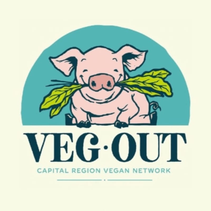 A smiling pig with a bunch of greens in its mouth sits in front of a teal semi-circle, with the words "VEG-OUT CAPITAL REGION VEGAN NETWORK" below.