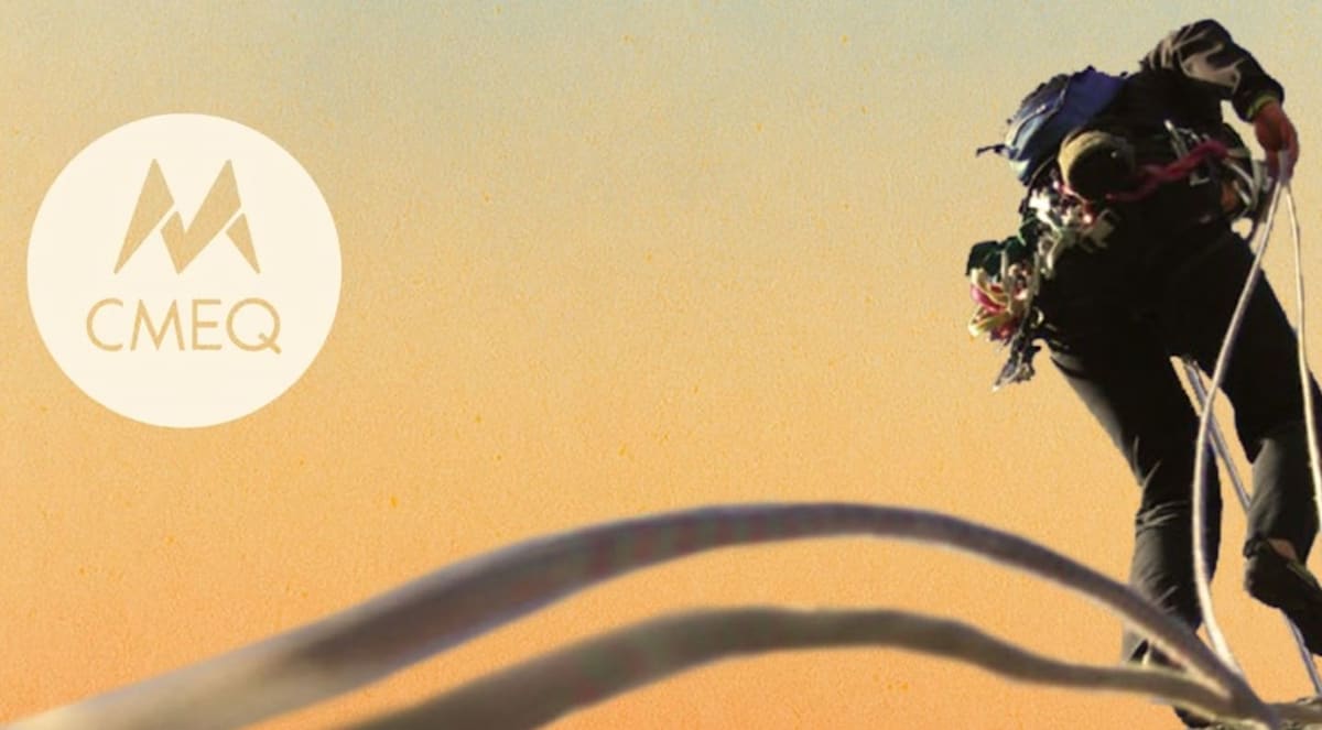 Blurred climbing ropes occupy the foreground, while a climber ascends a rock face against a soft, golden-hued sky.