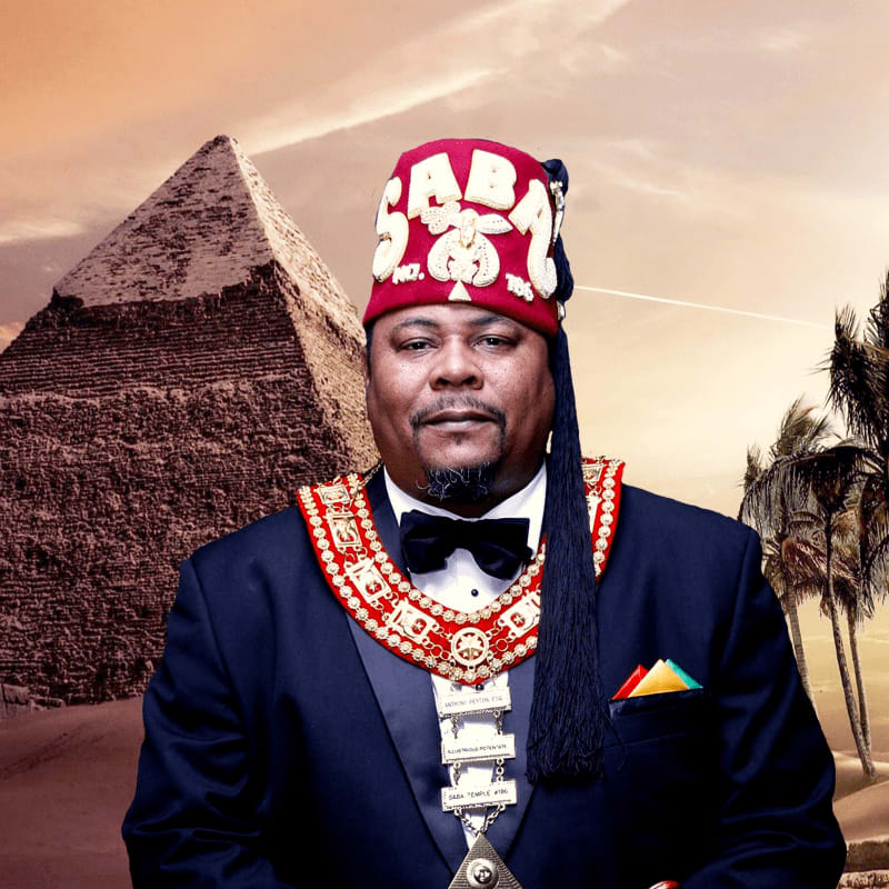 A man wearing a red fez and a formal suit with an ornate collar stands before a large pyramid and palm trees under a hazy sky.