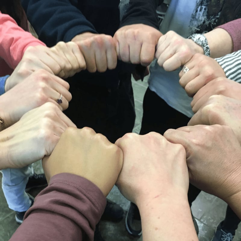 A circle of clenched fists joins together in the foreground against a background of people wearing casual clothing.