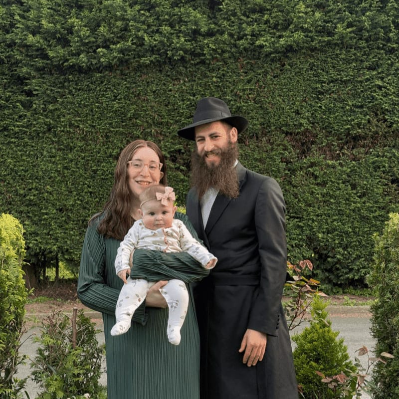 A family of three, a woman holding a baby, and a man in traditional attire, stand in front of a lush green hedge.