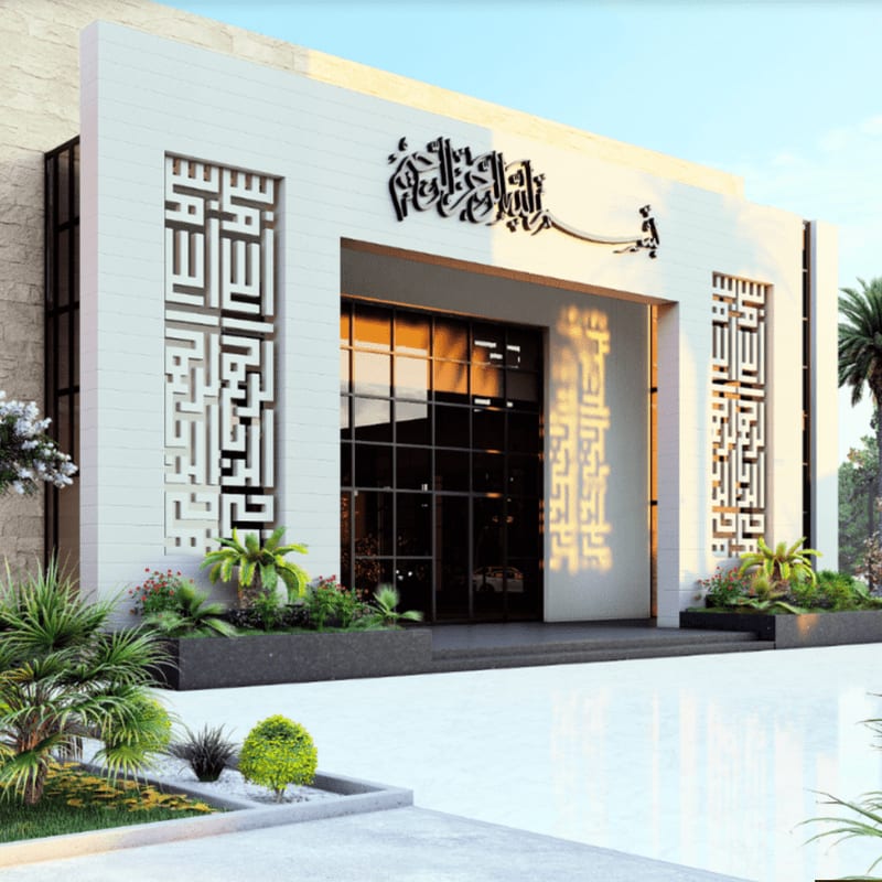 A modern white building with intricate geometric carvings and Arabic calligraphy stands behind a foreground of manicured greenery and a reflective white plaza.