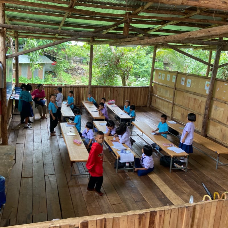 Children sit at wooden desks in an open-air classroom with a lush green forest in the background.