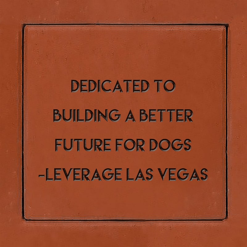 A square terracotta plaque with the text "DEDICATED TO BUILDING A BETTER FUTURE FOR DOGS -LEVERAGE LAS VEGAS" is set against a matching reddish-brown background.