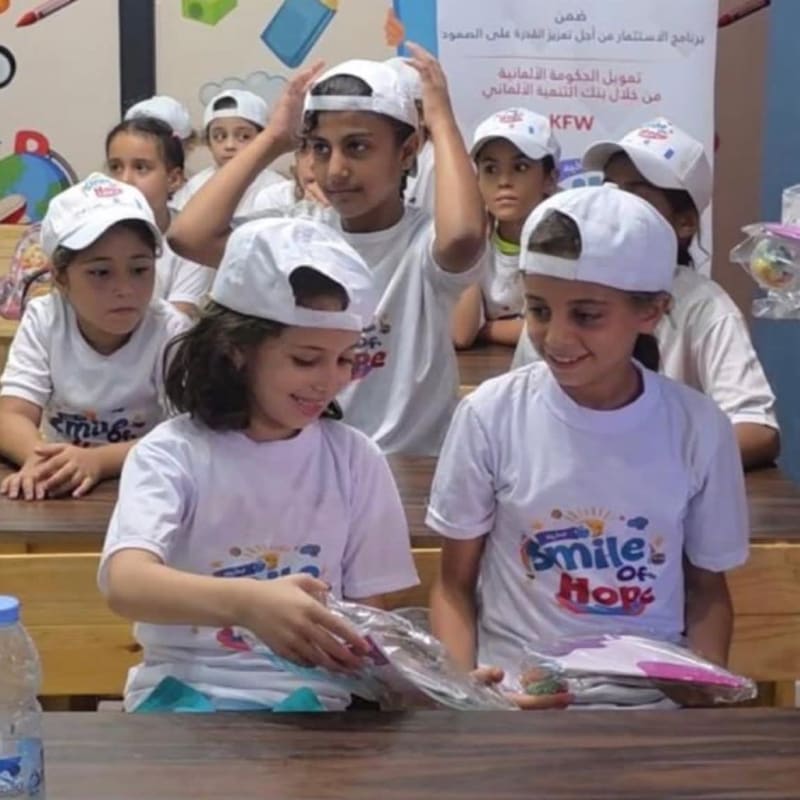Two young girls in white t-shirts and caps smile at each other while holding items, with other children in similar attire in the background against a wall with Arabic text and colorful graphics.