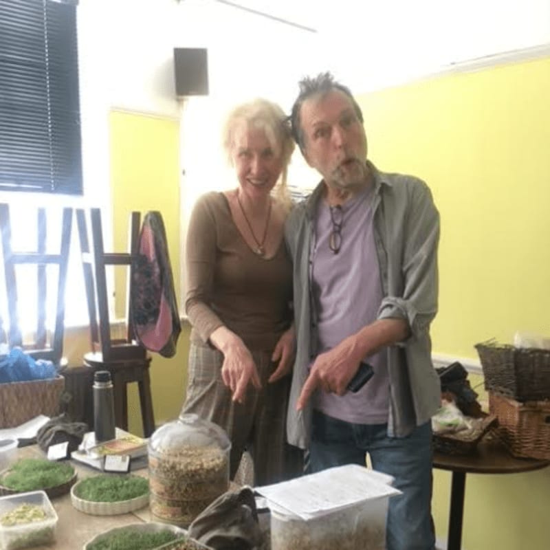 A man and a woman stand behind a table filled with containers of sprouts in a room with yellow walls.