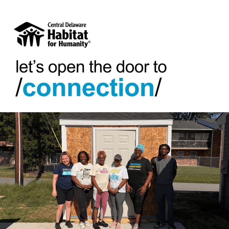 A group of six people stand in front of a small, newly constructed house with a residential building and trees in the background, all positioned below a Habitat for Humanity advertisement.