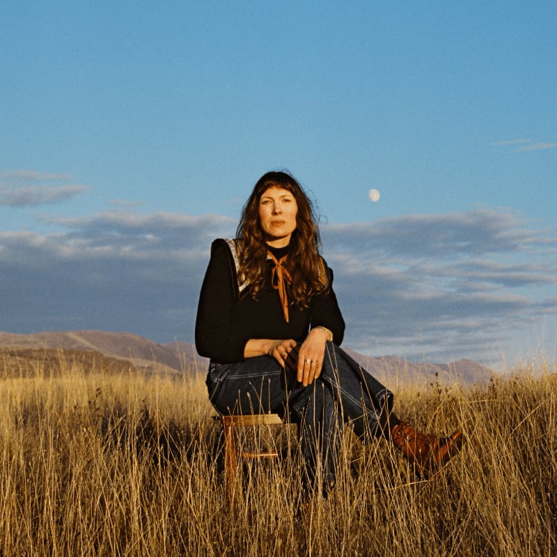 A woman sits on a wooden chair in a field of golden grass, with rolling hills and a pale moon in the blue sky behind her.