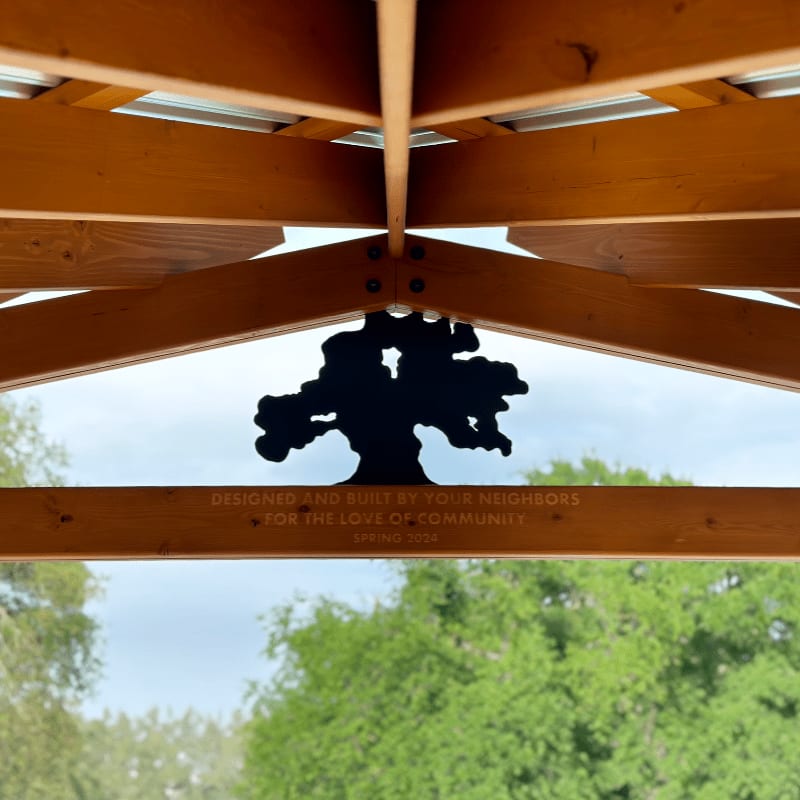 A black tree silhouette and commemorative text are mounted on a wooden pergola structure against a background of lush green trees.