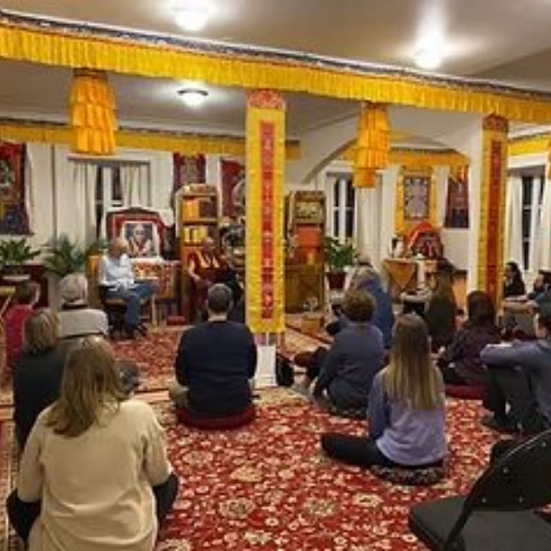 People sit on a red patterned carpet in the foreground, facing a teacher and ornate Buddhist altars in a room decorated with yellow fabric and traditional banners.