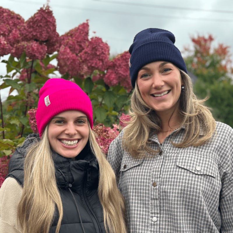 Two smiling women wearing beanies stand in the foreground against a background of pink flowering shrubs.