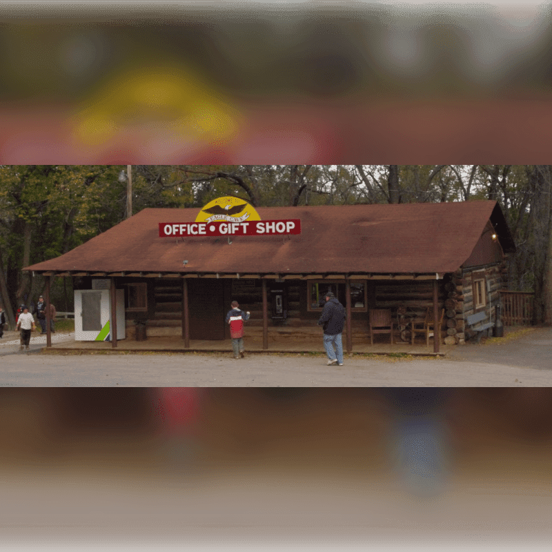 People stand in front of a log cabin office and gift shop set against a backdrop of autumn trees.