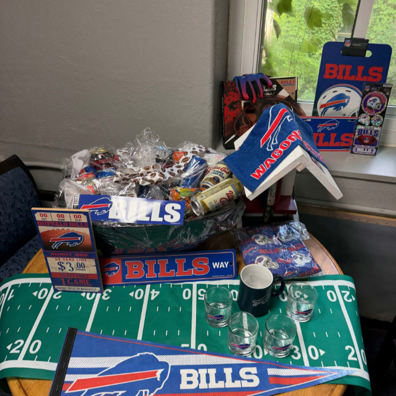A collection of Buffalo Bills football merchandise and snacks sits on a table in the foreground, with a window and green foliage visible in the background.