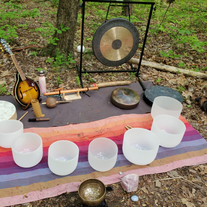 Various musical and sound healing instruments, including singing bowls and a gong, are arranged on blankets in a wooded forest setting.