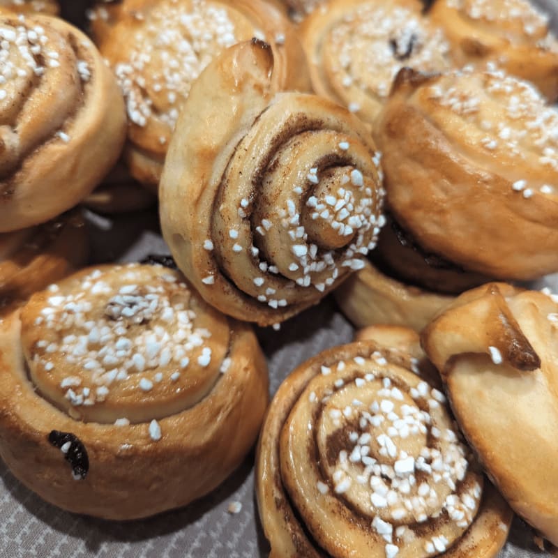 Golden-brown cinnamon rolls topped with pearl sugar fill the foreground and extend into a blurred background of more pastries.