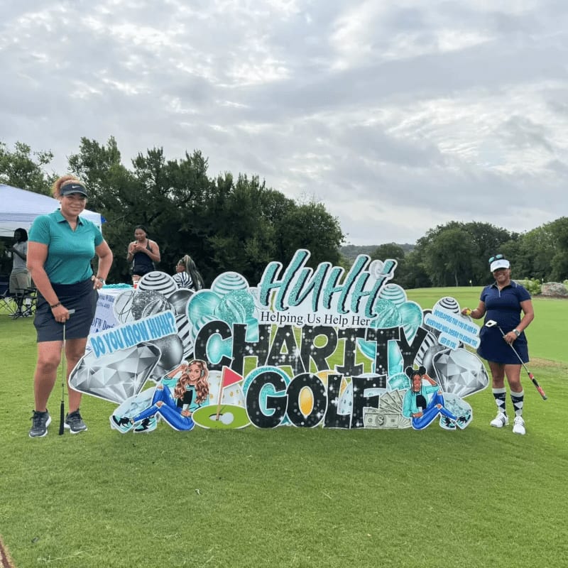 Two women stand on a green golf course beside a large, colorful "Charity Golf" sign under a cloudy sky.