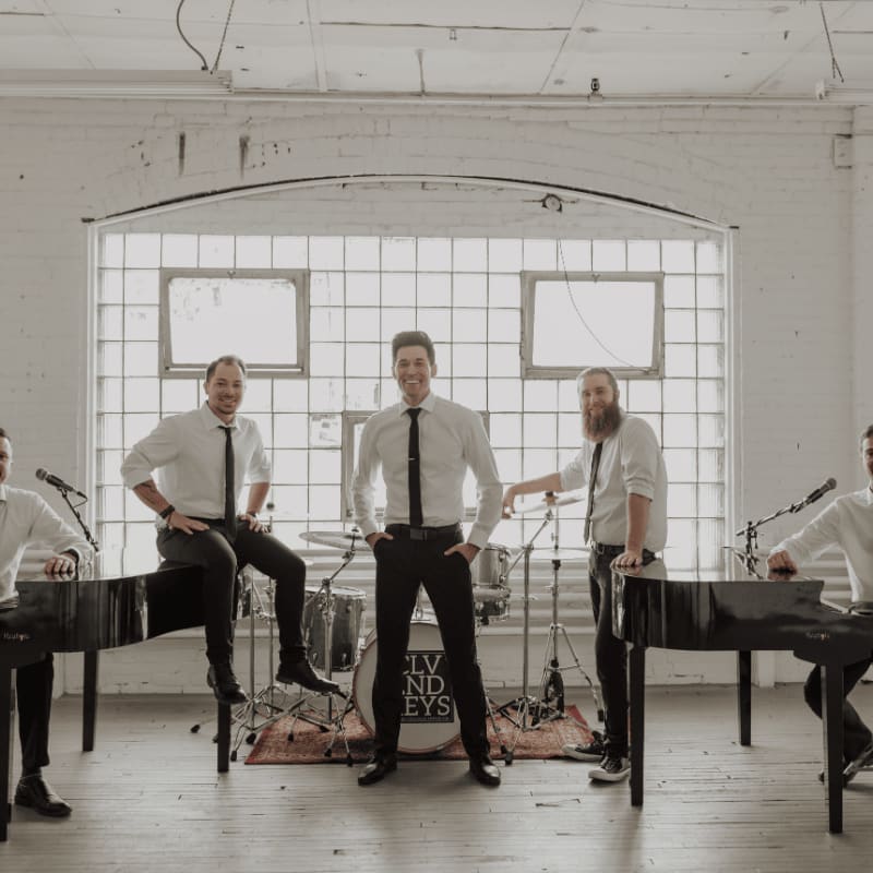 Five men in white shirts and black ties pose with musical instruments in a bright, white-walled room with a large grid window.