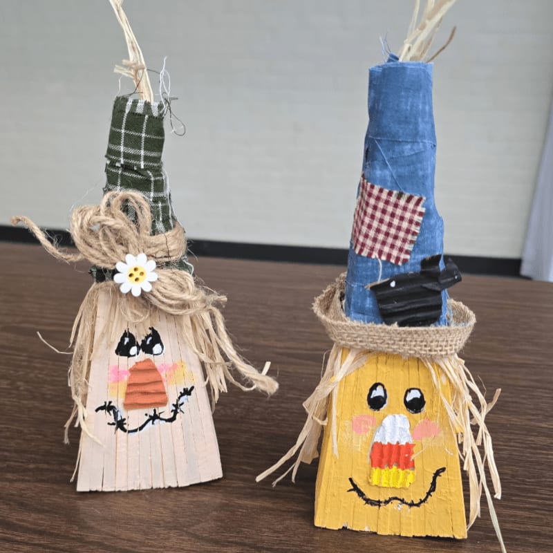 Two handmade scarecrow crafts with fabric hats stand on a dark wooden surface against a plain white wall.