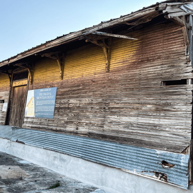 A weathered wooden building with a corrugated metal base and an informational sign stands against a clear blue sky.