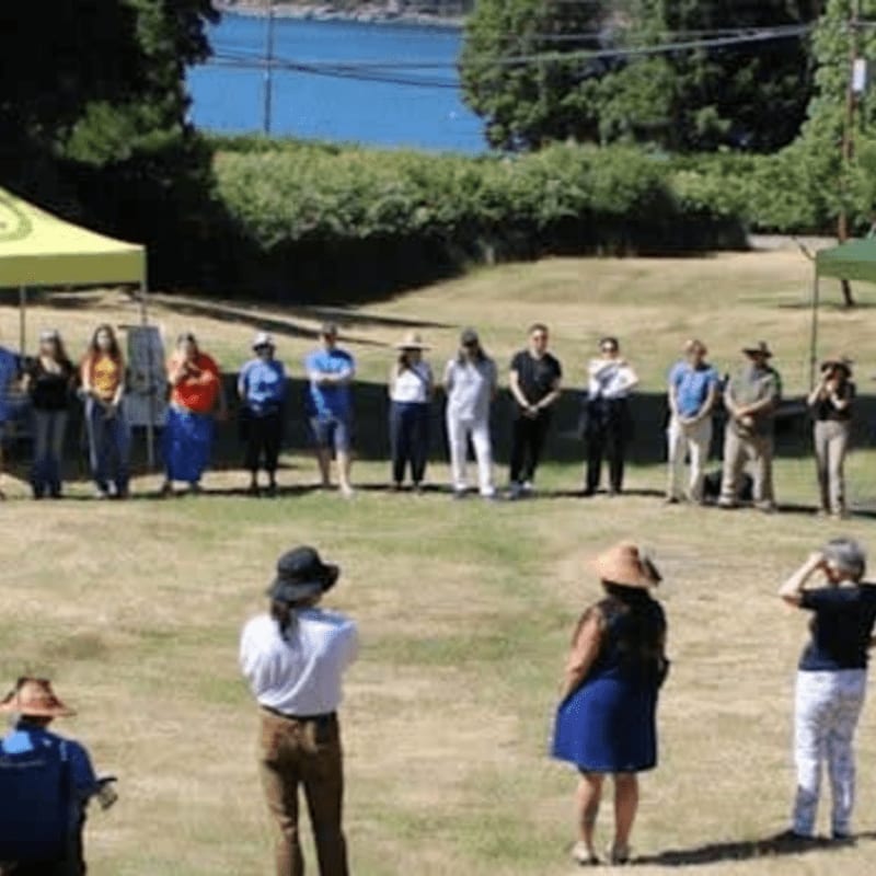 A group of people stand in a circle on a grassy field with yellow tents and a distant body of water in the background.
