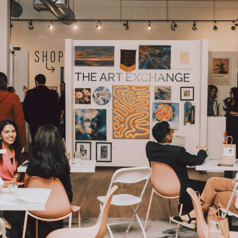 People sit and stand in a gallery space in the foreground, while a white wall displaying various artworks under the sign "THE ART EXCHANGE" fills the background.