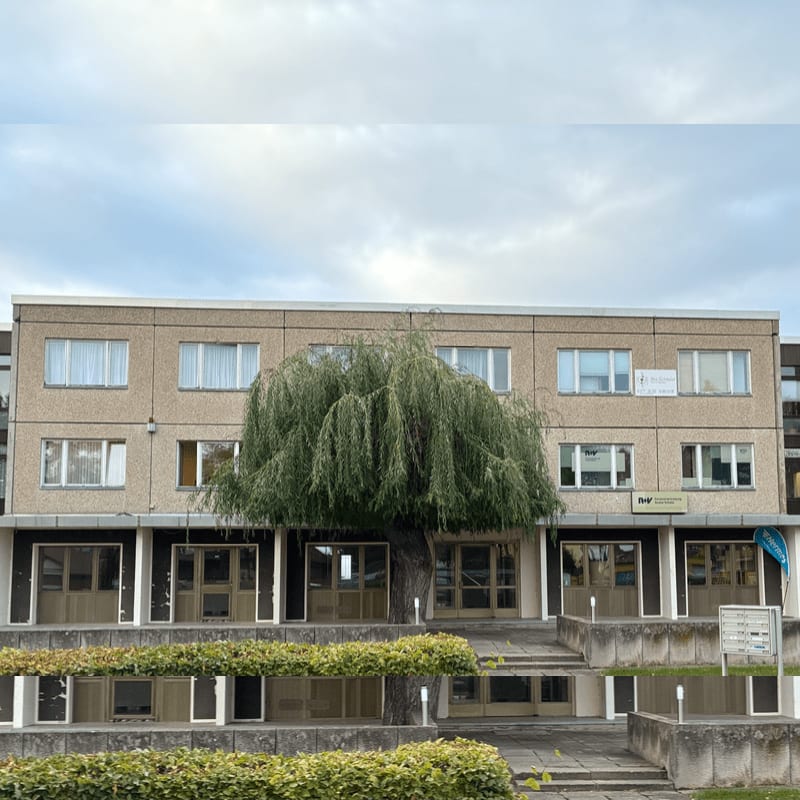 A large weeping willow tree stands in the foreground before a two-story beige office building under a cloudy sky.