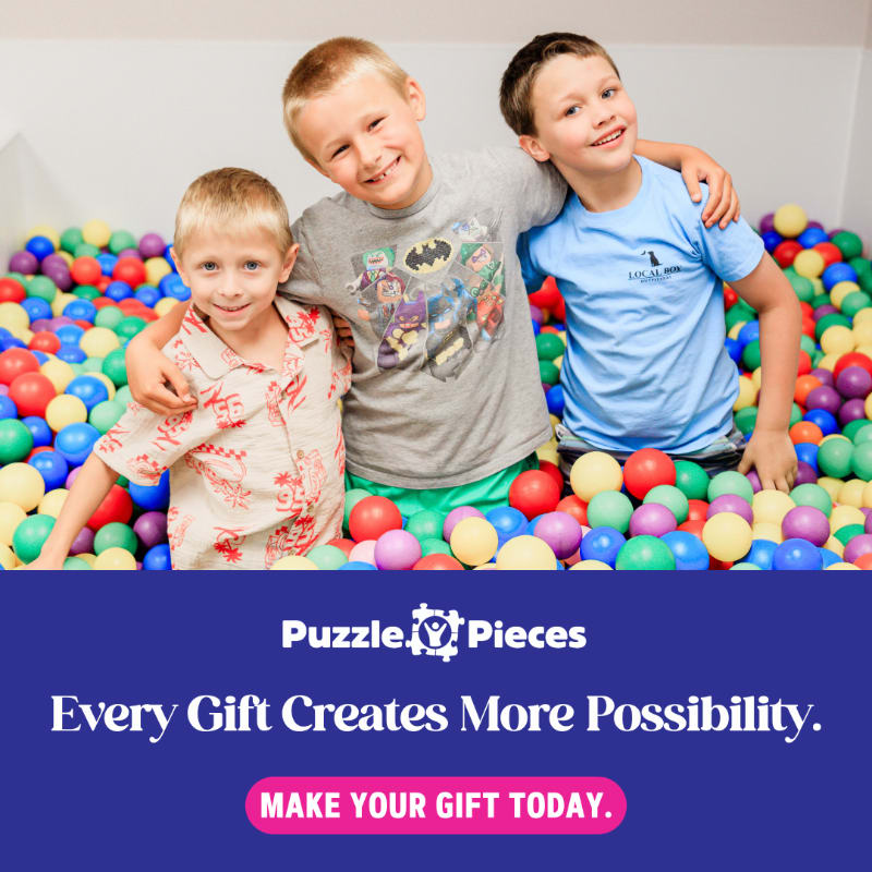 Three smiling young boys sit together in a colorful ball pit against a plain white background.