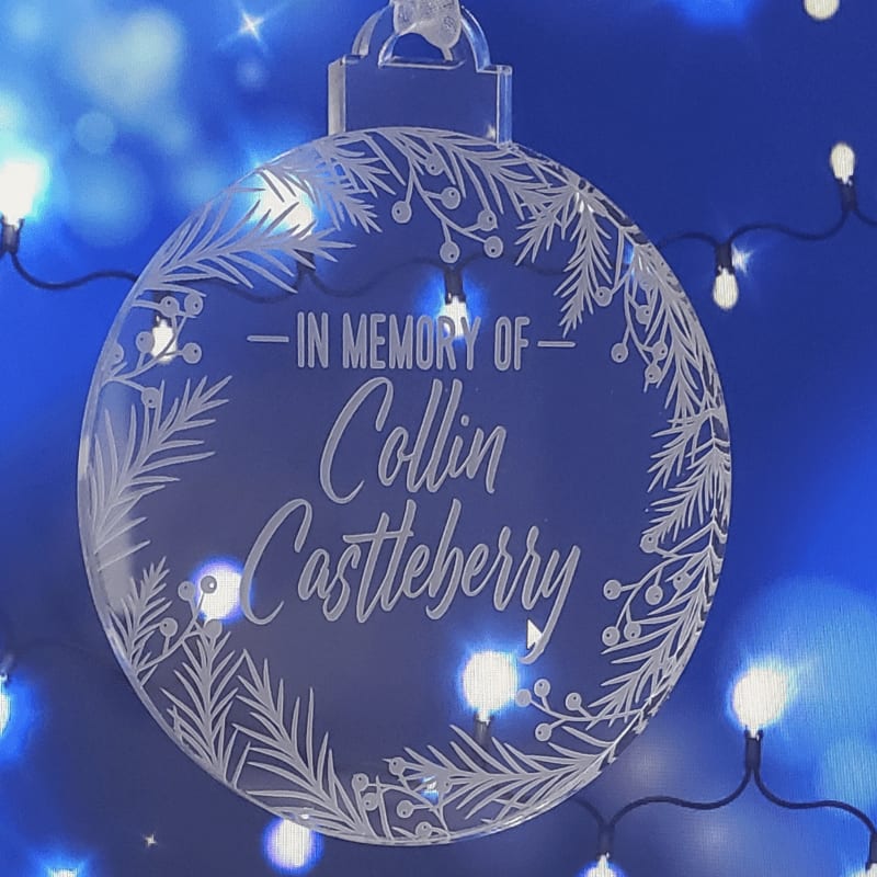 A clear circular ornament with "Emily & Jhon married 01.01.2023" etched on it hangs from a Christmas tree, with blurred green branches in the foreground and background.