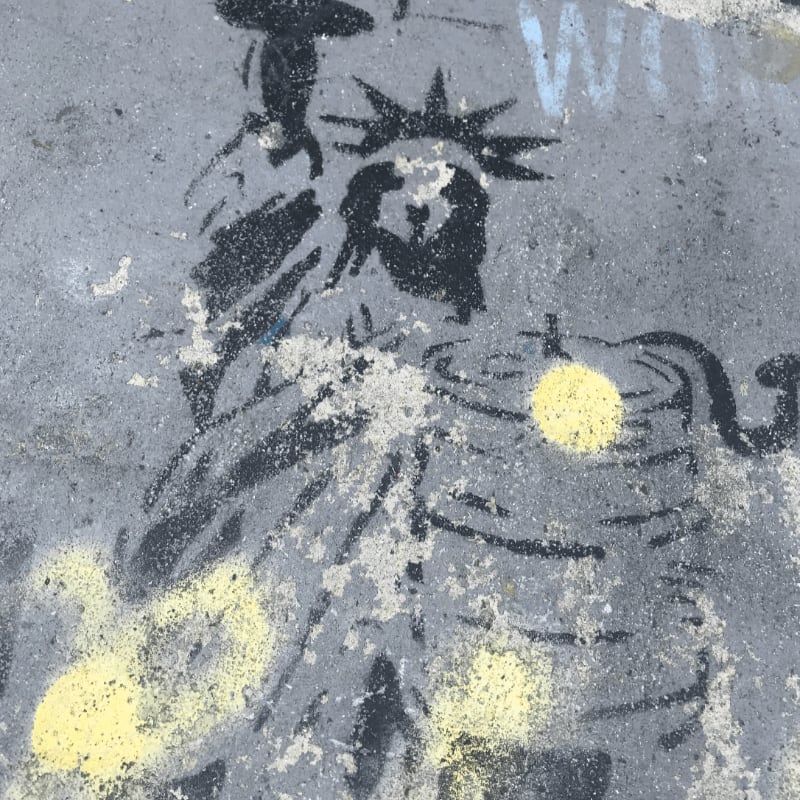 A black stencil of the Statue of Liberty is spray-painted onto a textured gray concrete surface with yellow paint splatters.