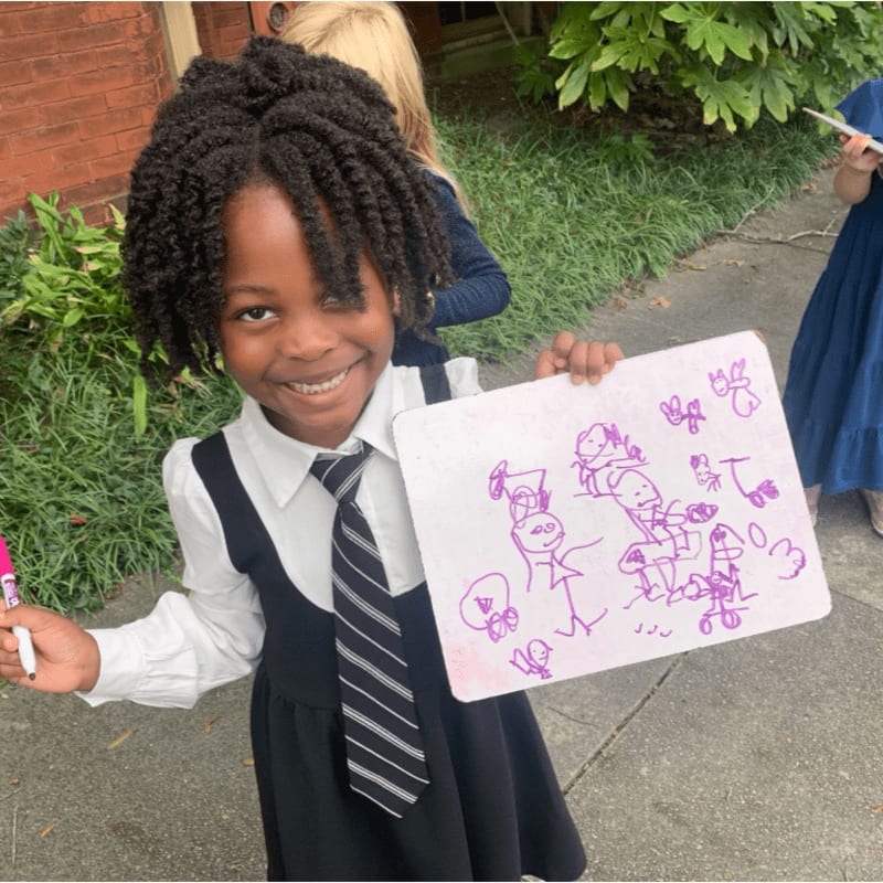 A smiling young girl in a school uniform holds up a drawing in front of a brick building and green foliage.