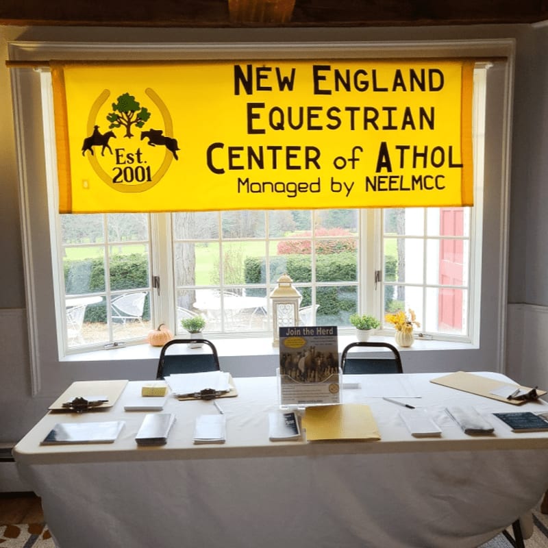 A white table covered in papers and brochures sits in the foreground, while a yellow banner for the New England Equestrian Center of Athol hangs in front of a window overlooking a green outdoor area.