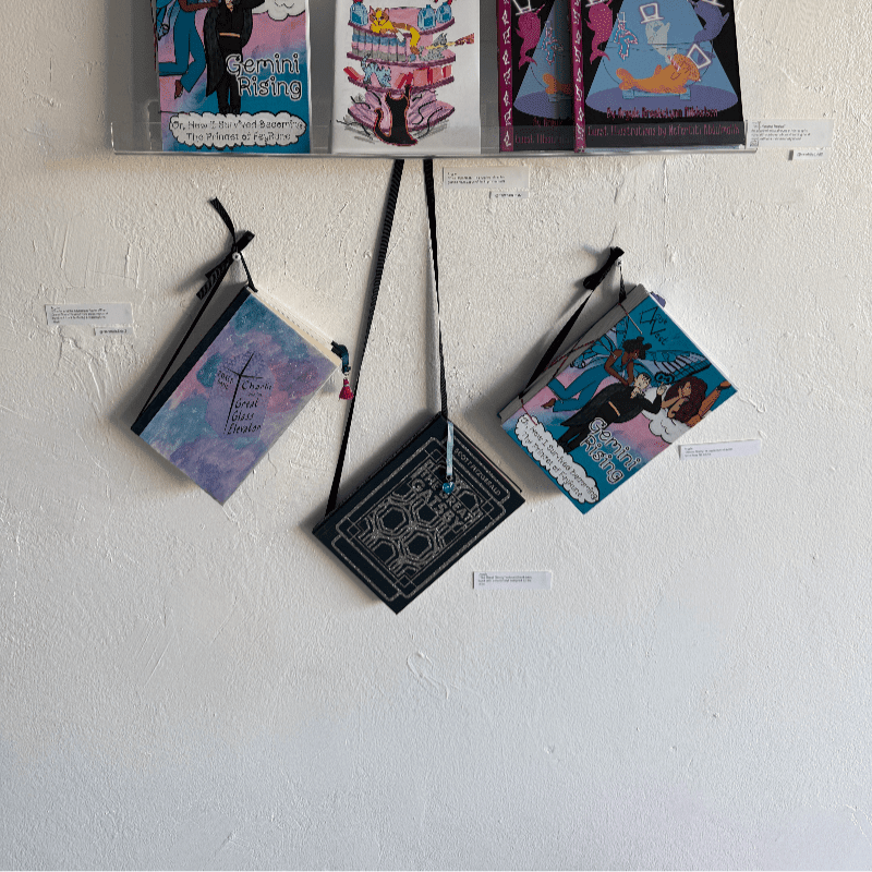 Three small art books hang from black ribbons against a plain white wall, with three more books displayed on a shelf above them.