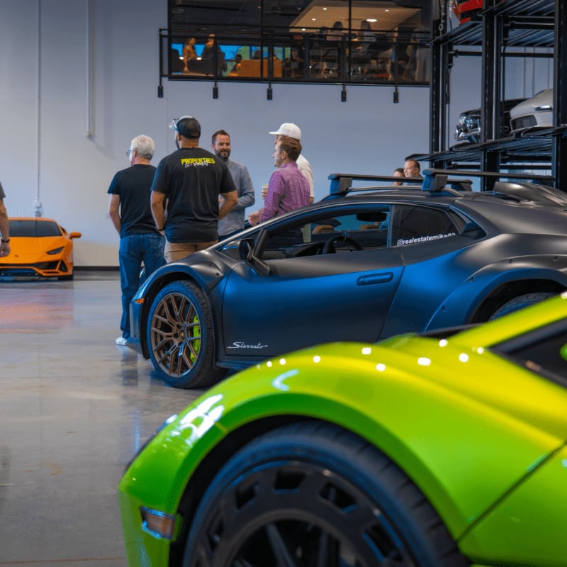 A bright green sports car sits in the foreground, with a matte black supercar and a group of men standing behind it in a modern garage.