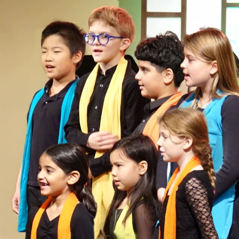 A group of children in black outfits with colorful scarves sing together against a plain, light-colored background.