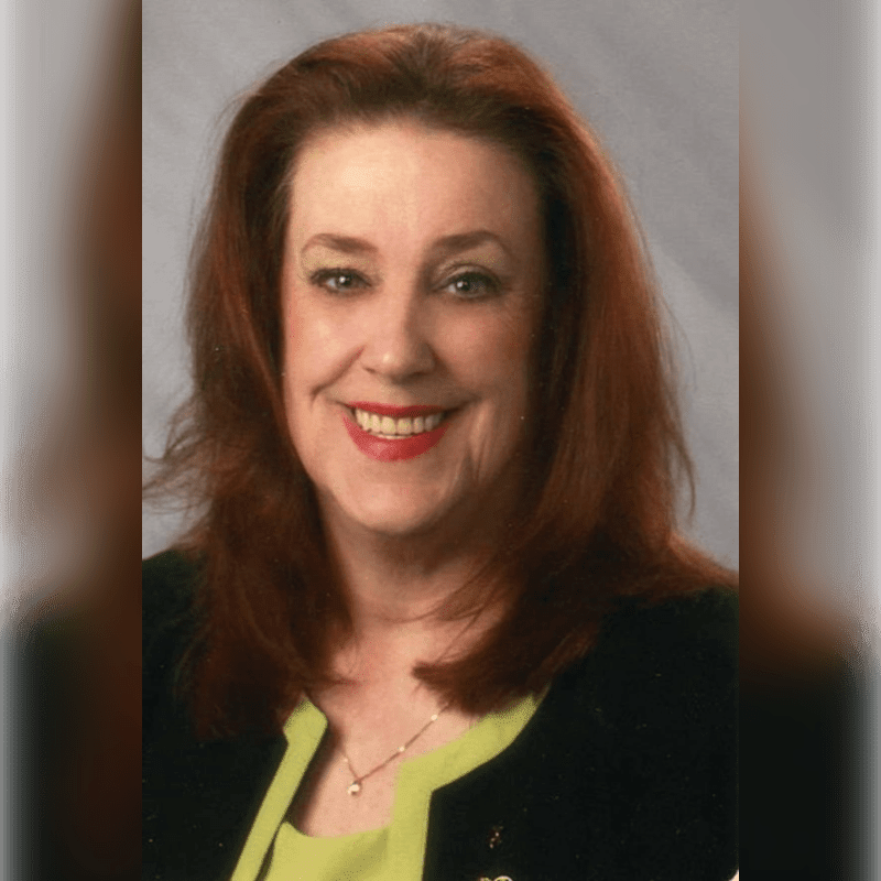 A smiling woman with red hair wearing a black blazer and lime green top stands against a plain grey background.