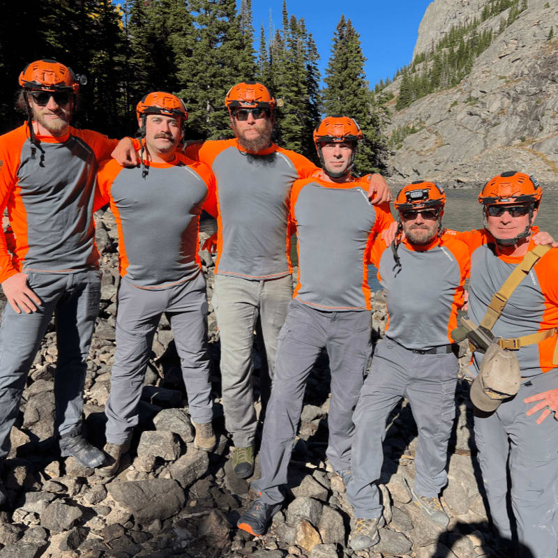 Six men wearing orange and grey outdoor gear and helmets stand together on a rocky shore with a forest and mountain in the background.